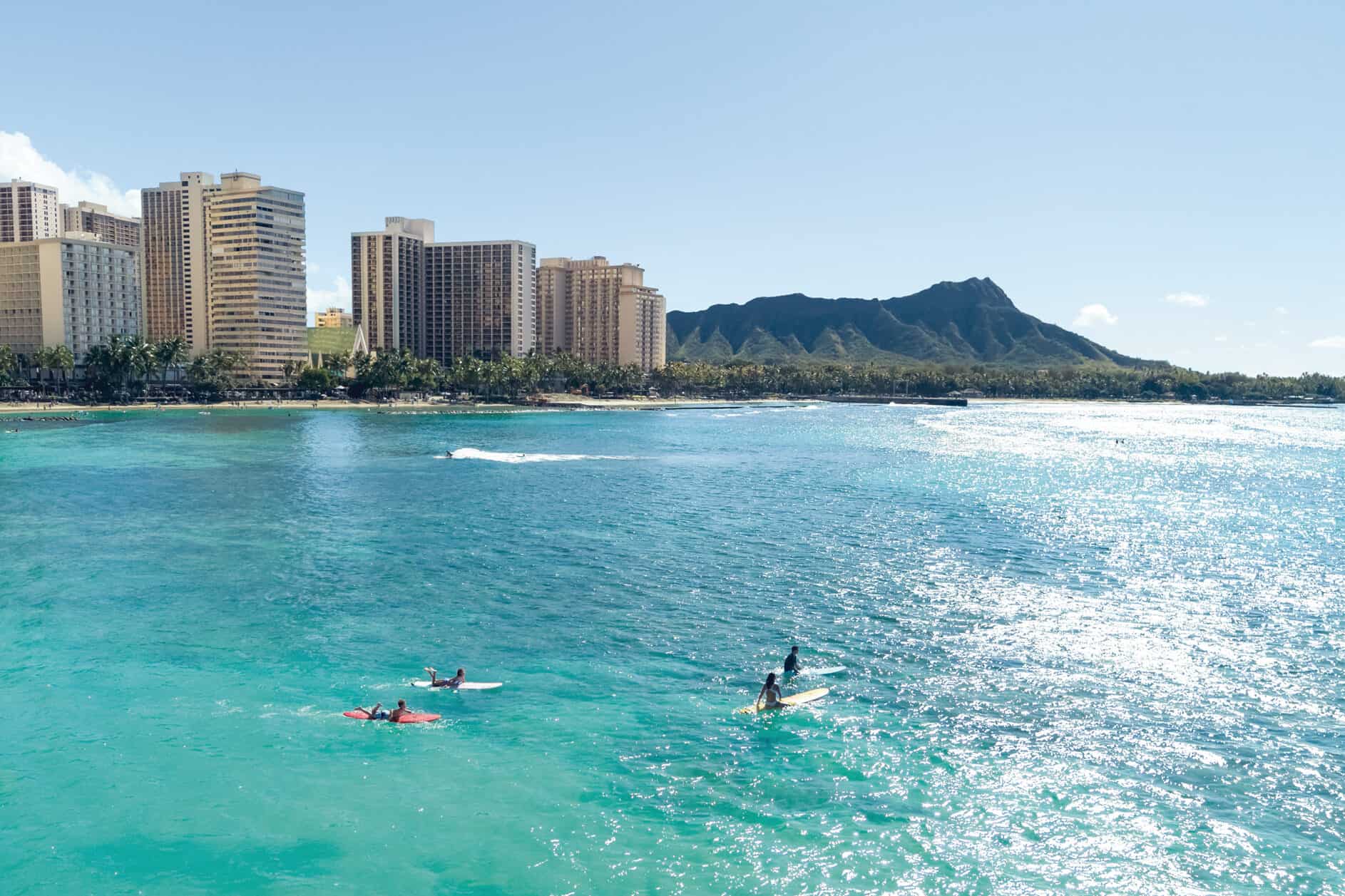 4 surfers in calm turquoise waters in front of a city with a mountain in the background