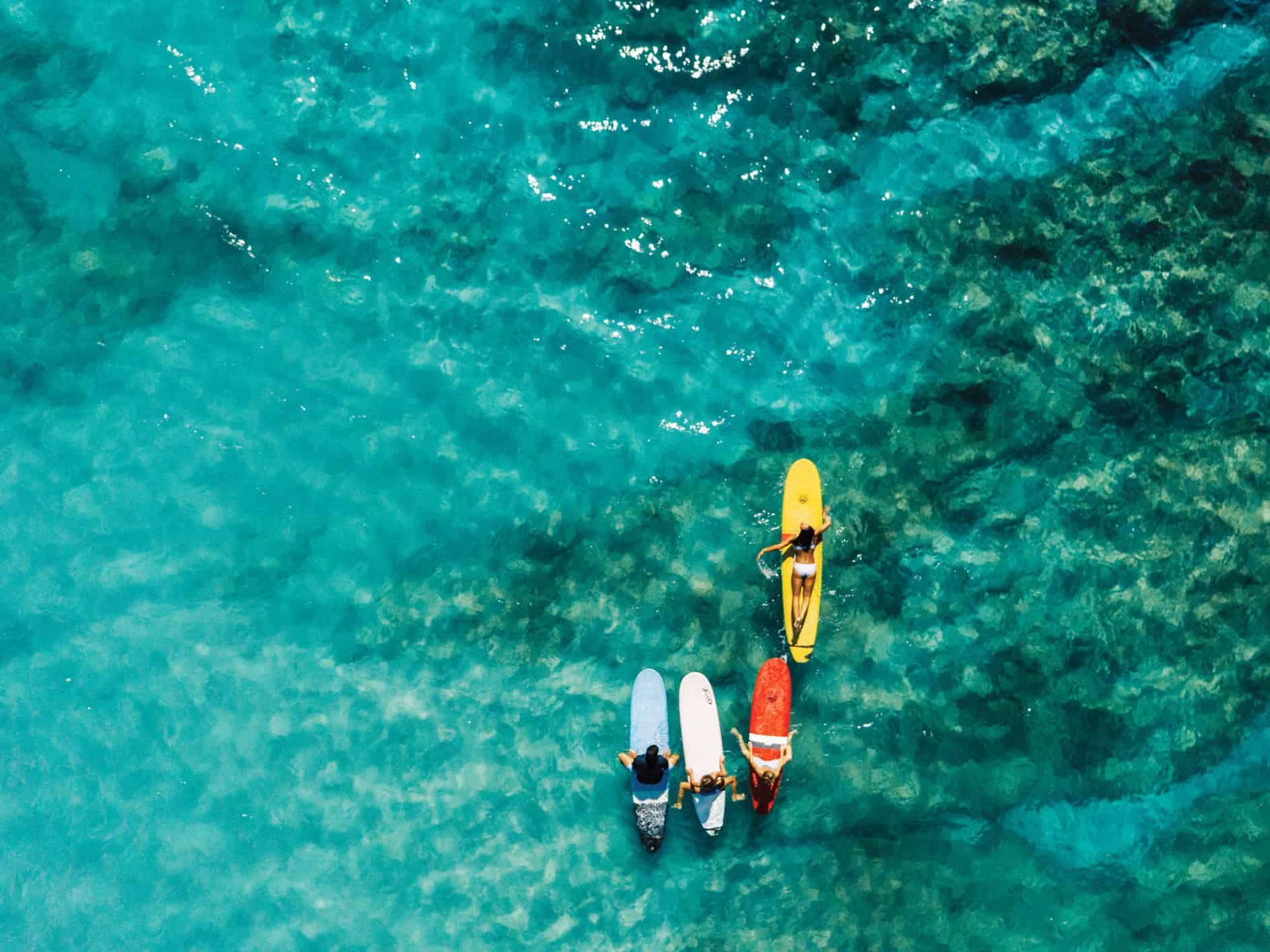 An overhead shot of a bright turquoise ocean, and blue, white, red and yellow surfboards in the water