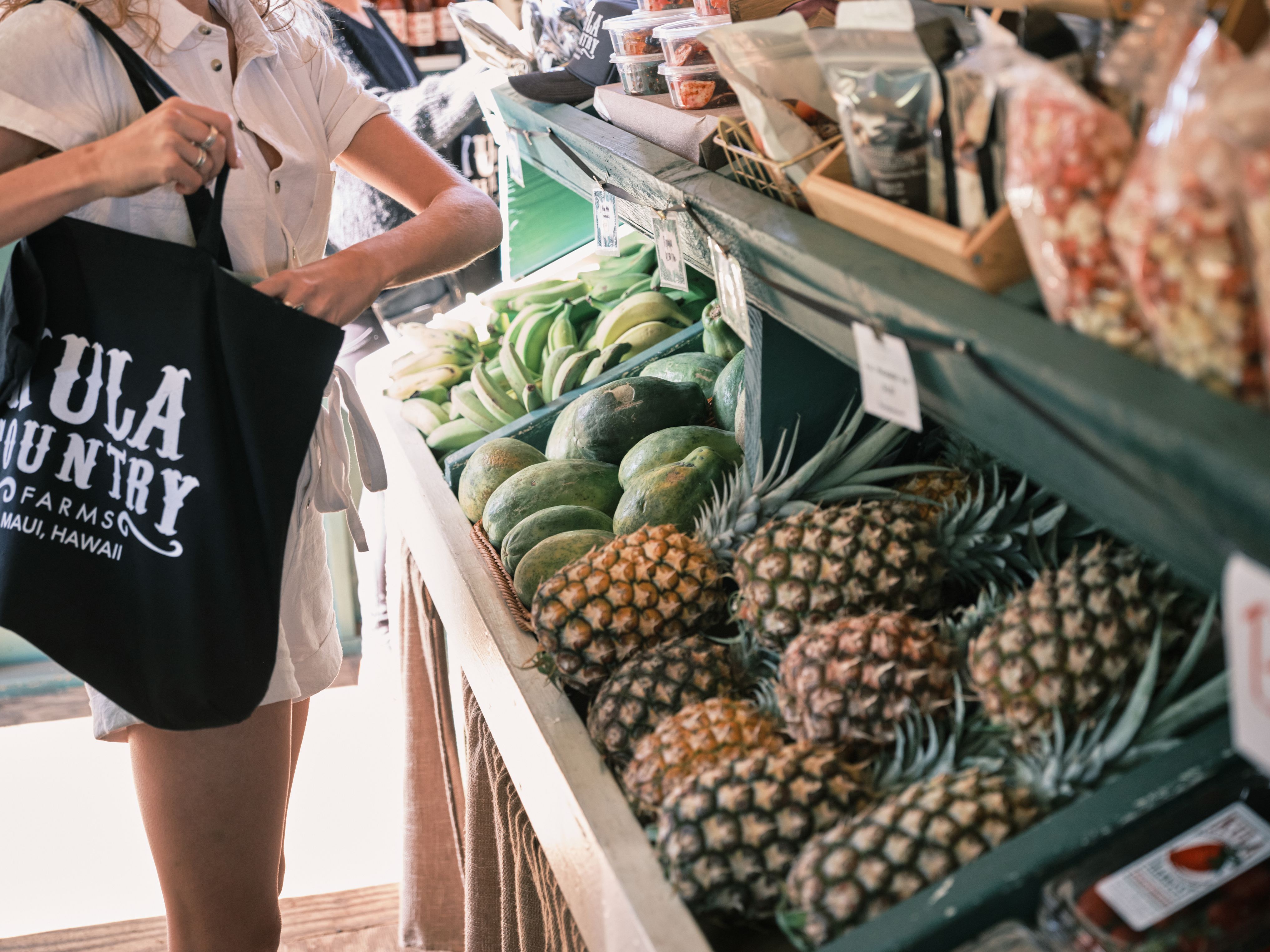 A woman holding a black shopping bag shopping through a row of pineapples at a farm stand