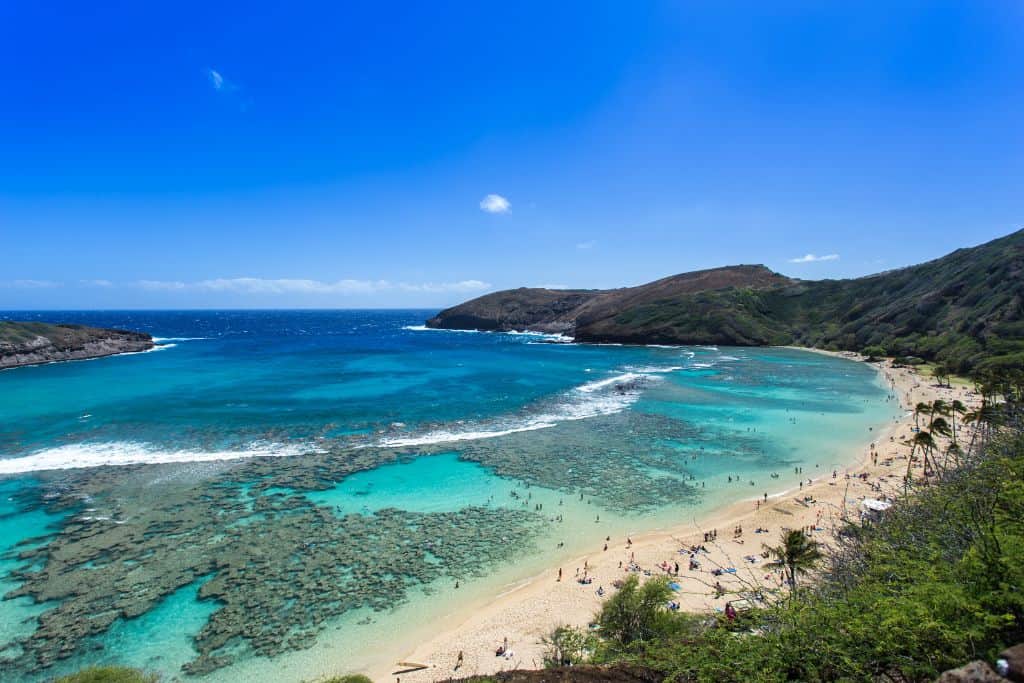 An aerial shot looking over a crowded beach with calm waters that are so clear you can see the reefs and a mountain in the background.