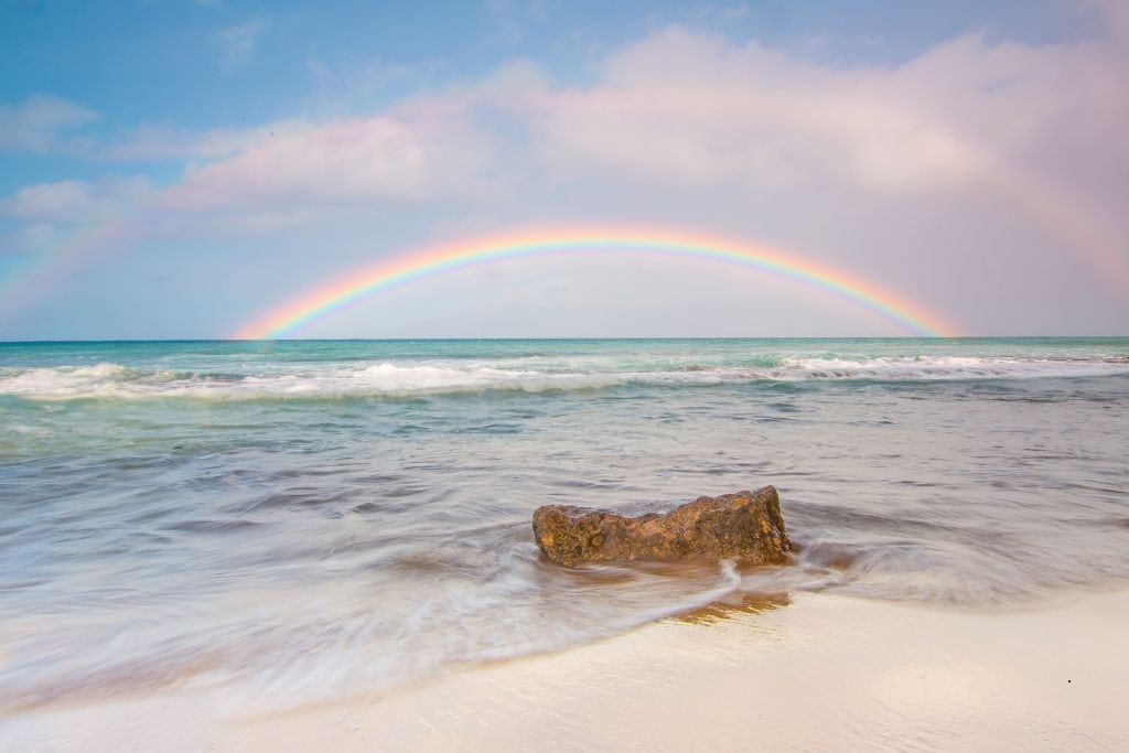 A white beach with a brown rock just at the waters edge and a rainbow in the background of a light turquoise colored ocean.