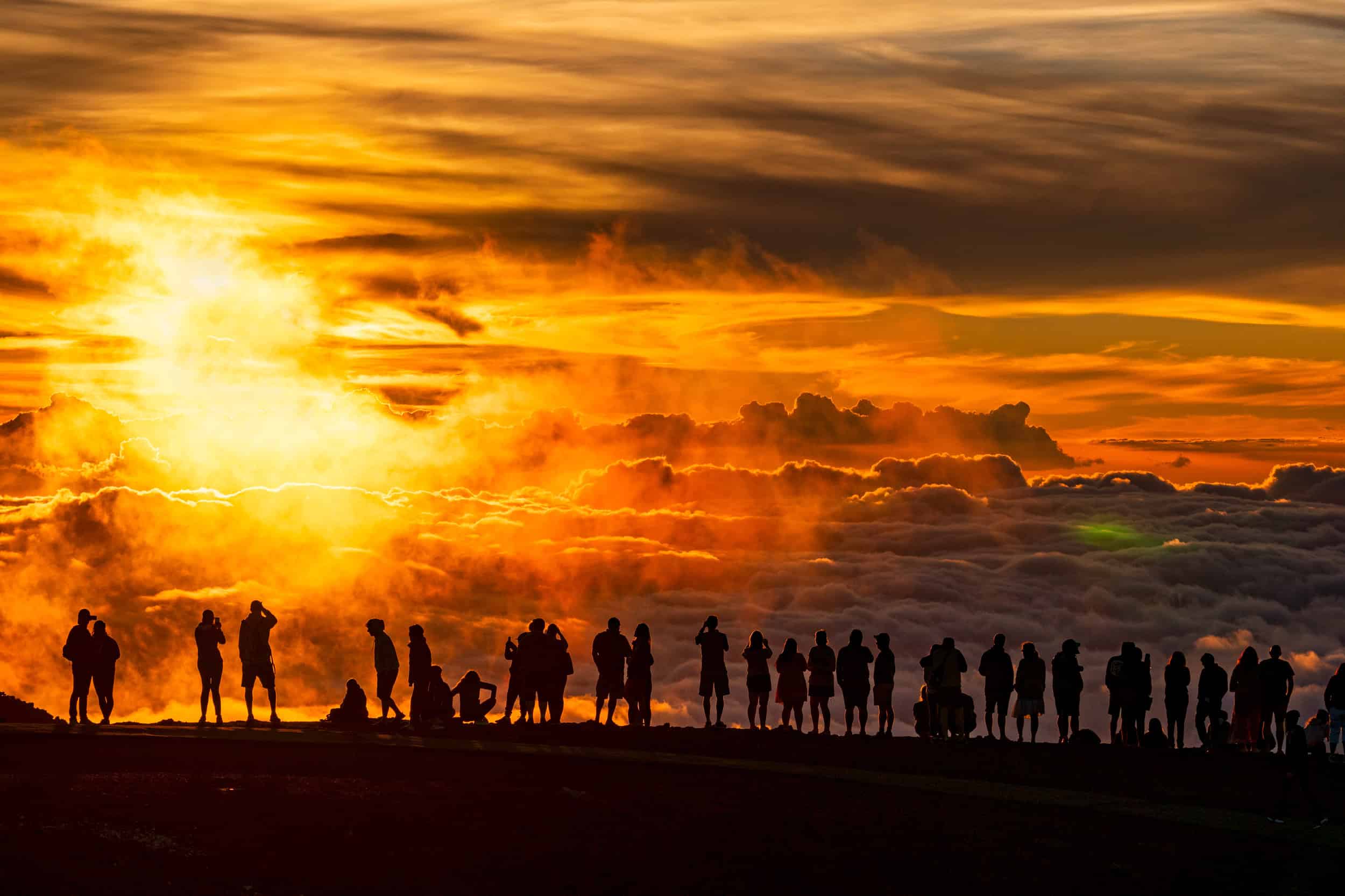 A bright orange sunset with a large yellow sun to the left and the shadows of a row of people on the edge of a mountain looking down at the sunset and clouds