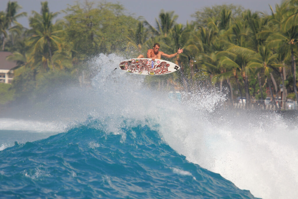 A surfer popping up high above an ocean wave with palm trees behind him