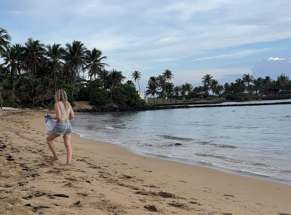 A blonde woman in a gray top and jean shorts walking on the beach carrying a bag to clean up garbage with palm trees in the background.