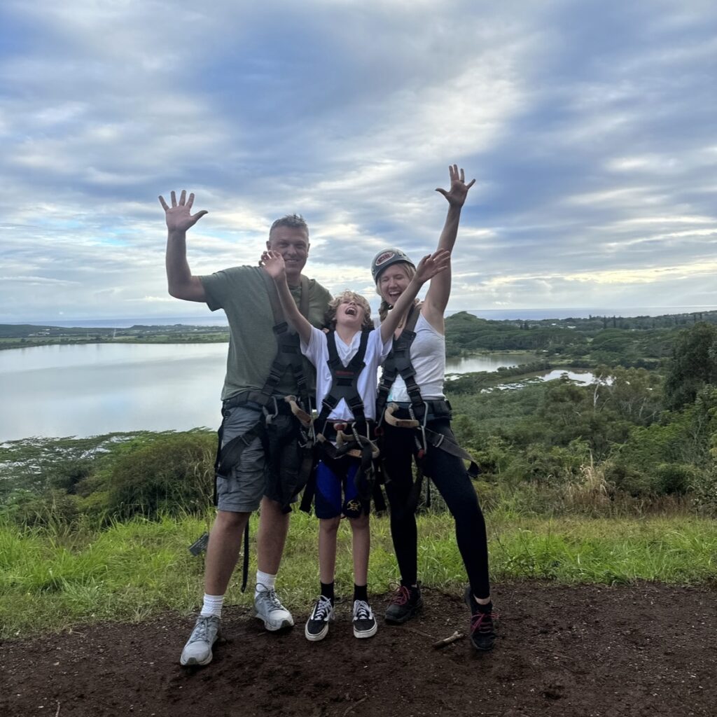 A man, child and woman wearing helmets standing in front of a tropical landscape putting their hands up in the air