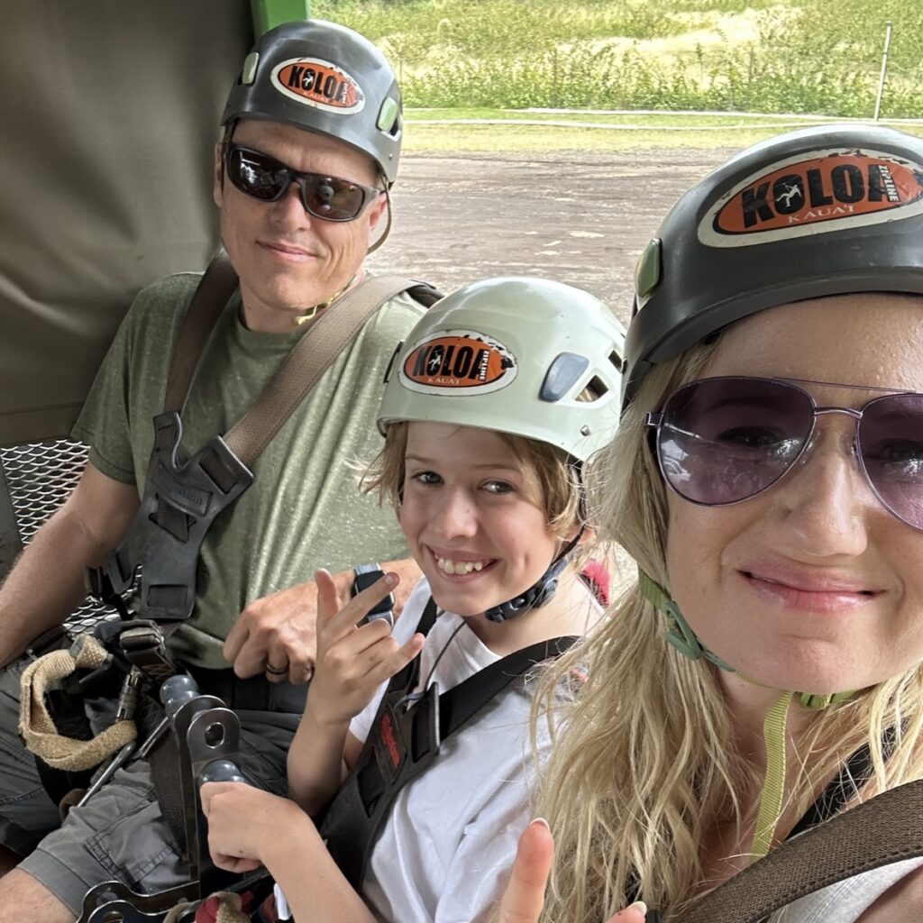 A smiling man in a green shirt and helmet sitting next to a child in a white shirt and helmet and a blonde woman smiling in a helmet