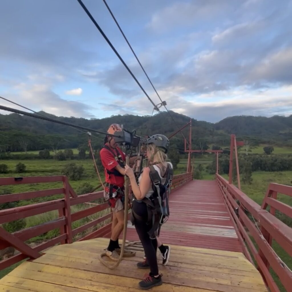 a man in a red shirt and a helmet helping two zipliners wearing helmets get unclipped after ziplining over a tropical jungle