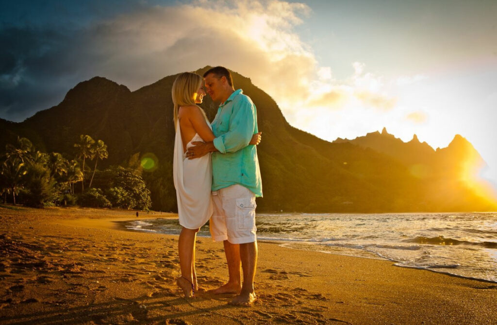 A blonde woman in a white dress and a man in a teal shirt and white shorts embracing on a beach in front of a tropical mountain landscape