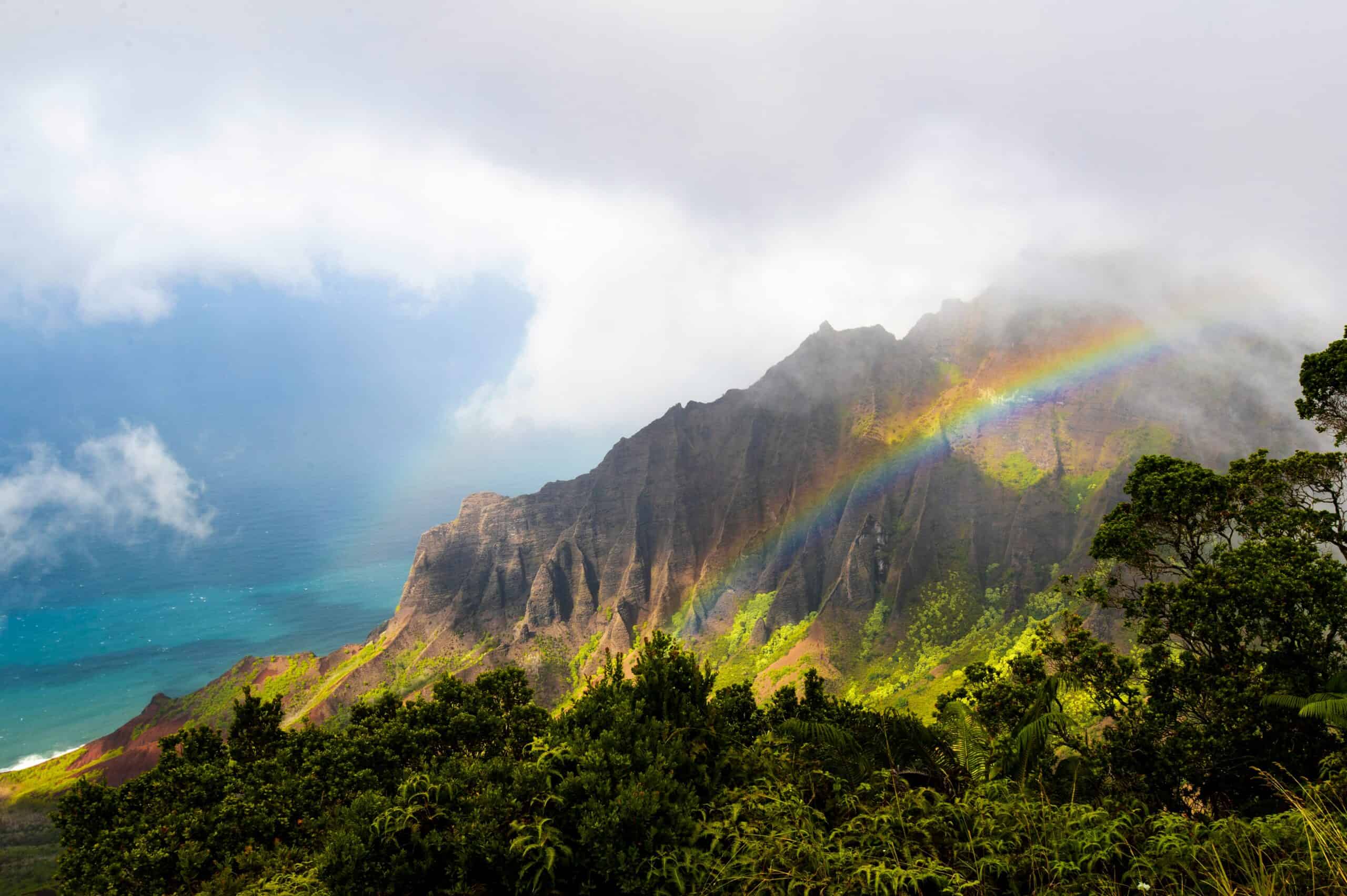 A lush green tropical mountain with a rainbow in the foreground and ocean in the background.