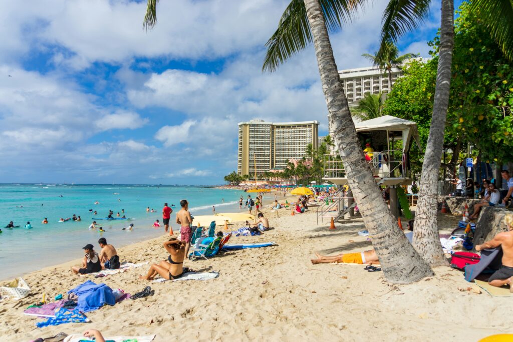 A crowded beach with a palm tree trunk in the foreground, people wearing colorful swim wear laying out on a beach and a city in the background