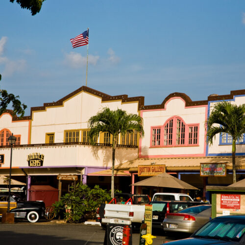 An old looking town with white buildings that have colorful trim and American flag off the trop. Older cars and people in the foreground.