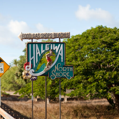 The side of the road with a guardrail and two signs, one that is yellow and says 35 MPH in black text against a white box and a blue town sign that says Food, Gas, Shops, Beaches with an arrow pointing to the left and Haleiwa North Shore with an image of a man surfing in red shorts