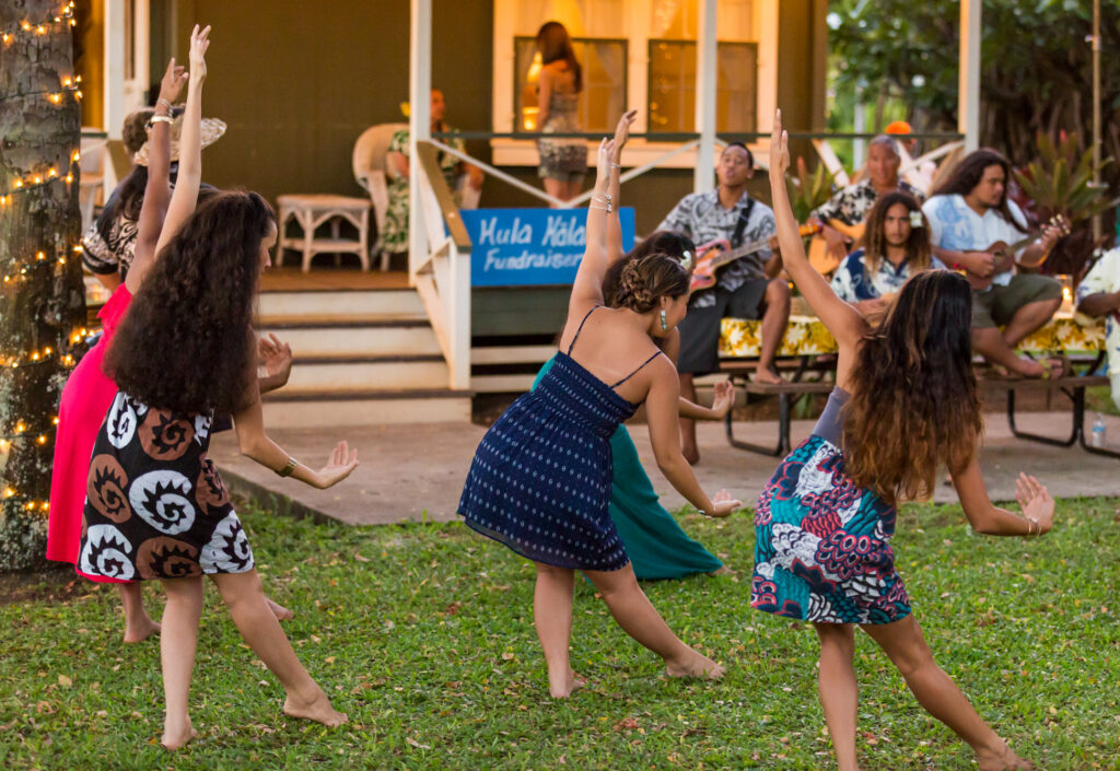 5 women in colorful shirts and dresses dancing hula with 4 musicians playing guitar  in the background