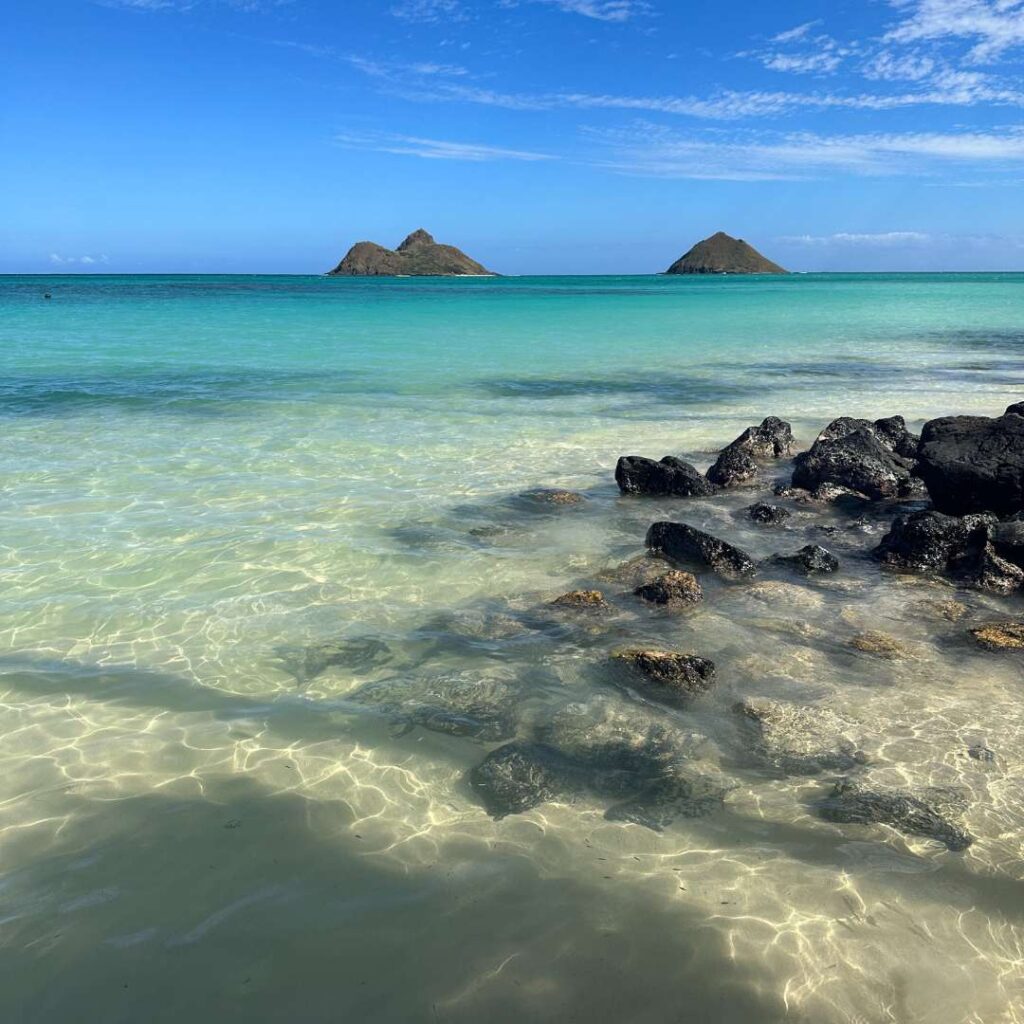 A calm turquoise ocean with rocks in the foreground and two islands in the background.