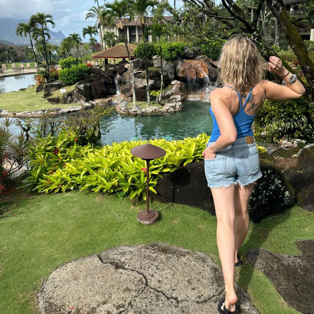 A blonde woman in jean shorts and a blue top looking over tropical pools and landscaping