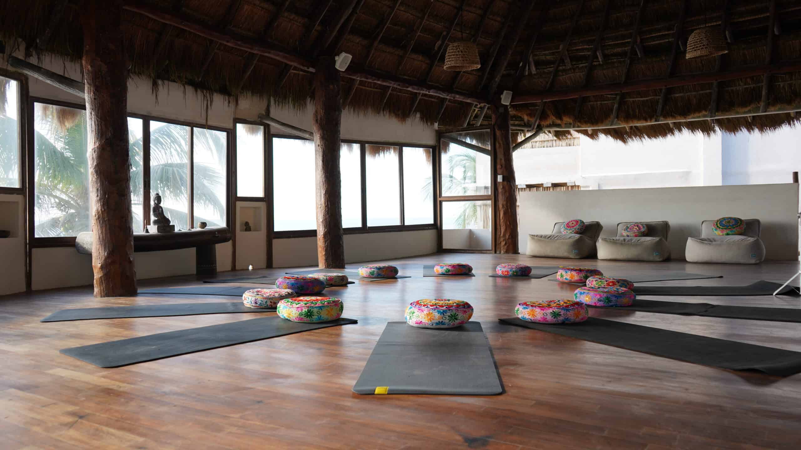 Yoga mats and bright colorful cushions set up in a circle in a room with many windows.