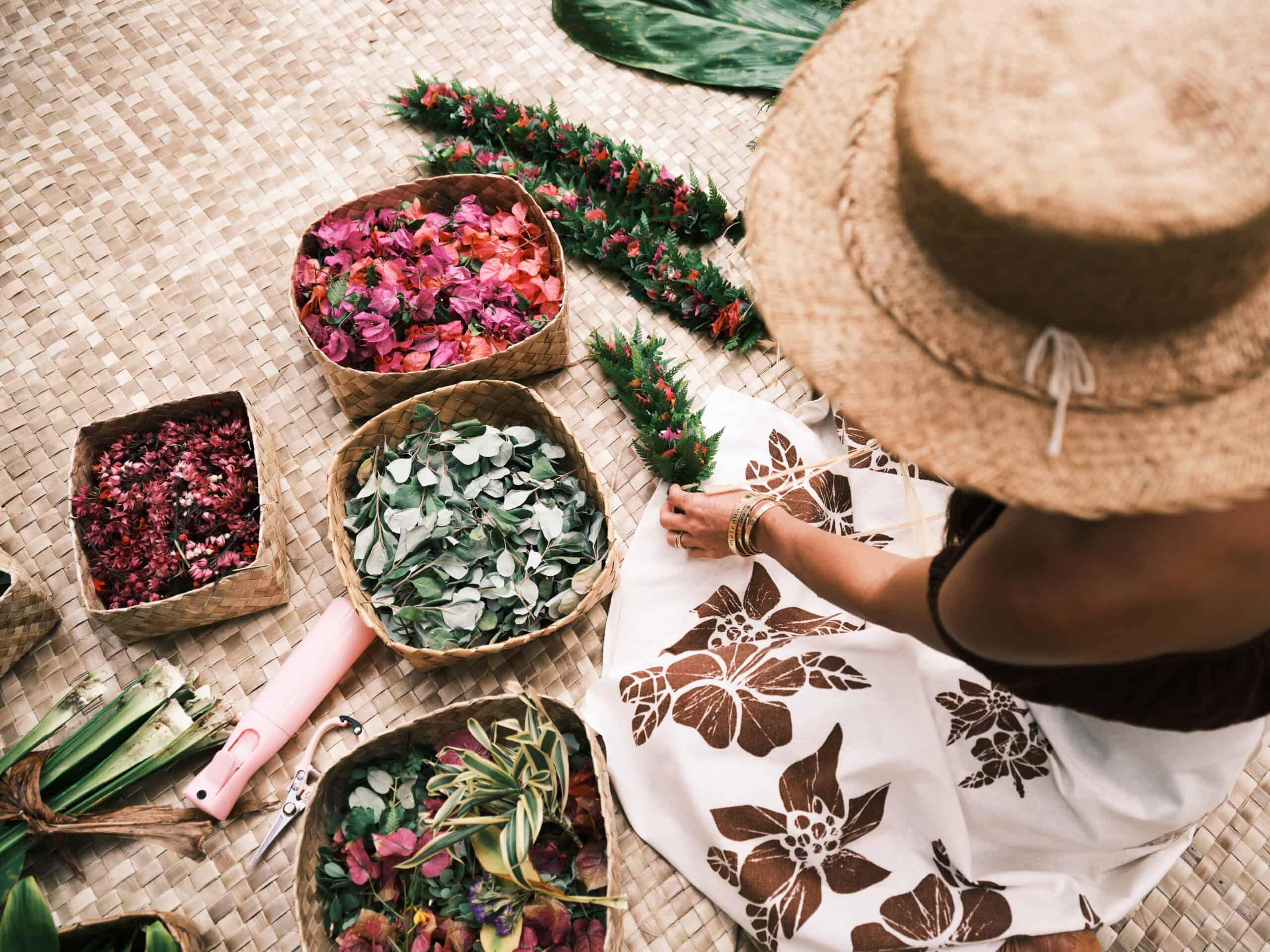 A woman in a white dress with brown flowers on it and a straw hat making Hawaiian lei's from baskets of flowers and greens