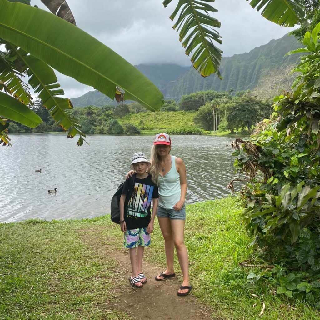 A boy and woman in a red hat standing in front of a lake with tropical mountains behidn them