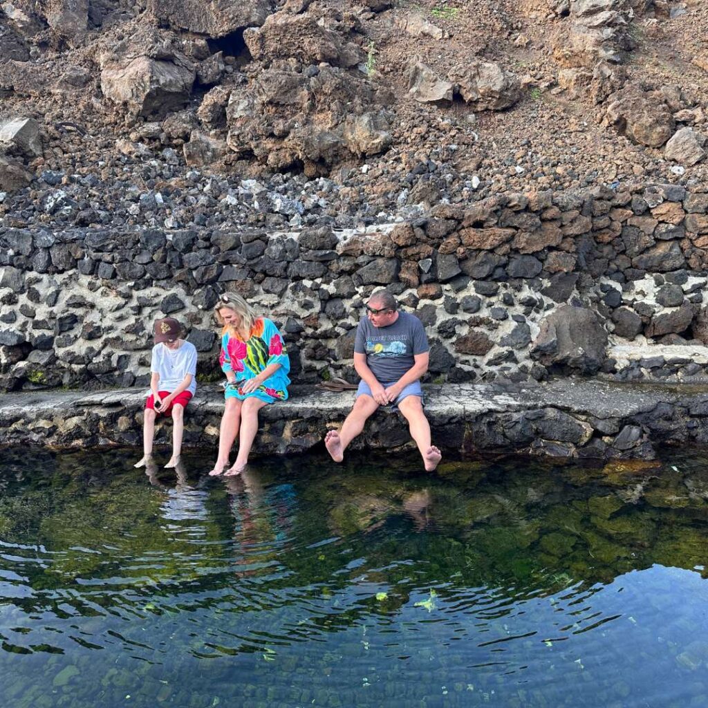 A boy, a woman, and a man dipping their feet in water with rocks behind them