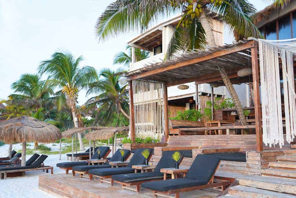 A white wooden building with palm trees around it and a row of black lounge chairs in the sand in front of it.