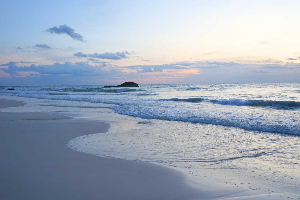 Ocean waves on the sand with a sunrise behind a rock.