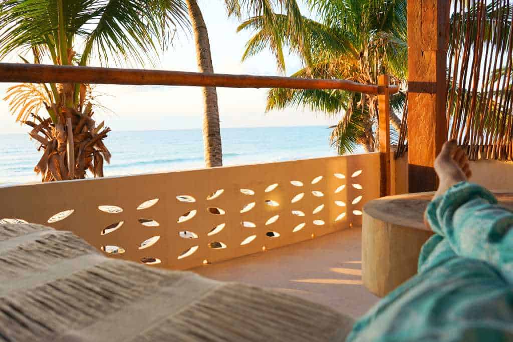 A sun filled patio looking at a palm tree and an ocean with a woman's legs relaxing on a table.