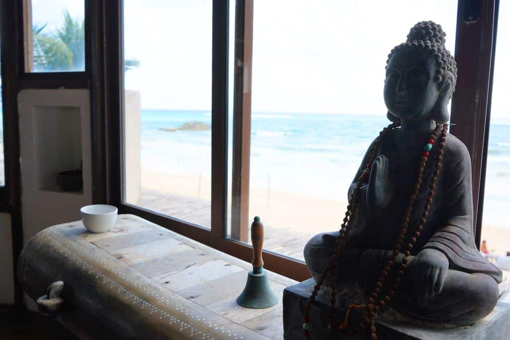 A buddha statue and hand bell on a table with an ocean out the window.