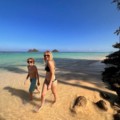 A blonde boy and blonde woman in their bathing suits and sunglasses walking down the sand with two islands in the background