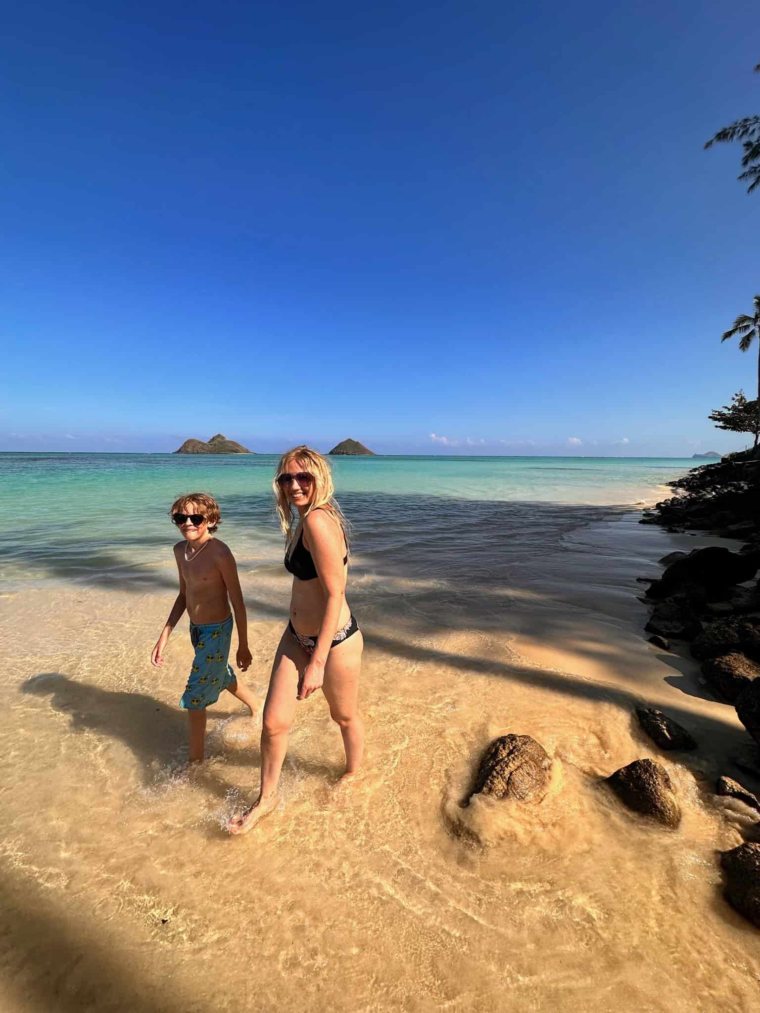 A blonde boy and blonde woman in their bathing suits and sunglasses walking down the sand with two islands in the background