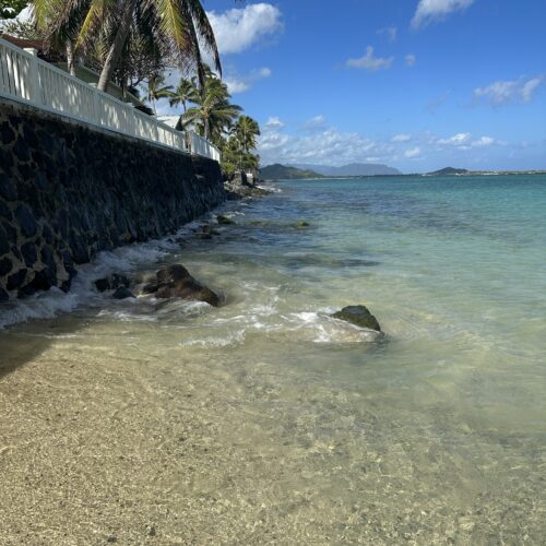 A stone and white retaining wall with palm trees above it to the left and a calm clear ocean with islands in the background to the right