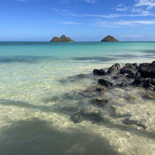 A clear turquoise calm ocean with rocks in the foreground and two triangular islands in the background