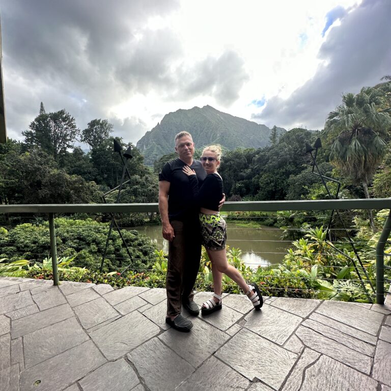 A man in black and a blonde woman in a black shirt and a green skirt with a tropical mountain, lake and trees in the background