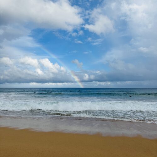 A yellow sandy beach with turquoise waves splashing on it, surfers in the water and a rainbow in the sky in the background