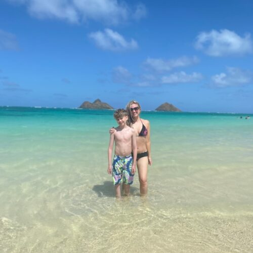 A blonde boy and woman in their bathing suits standing in the ocean with two islands behind them