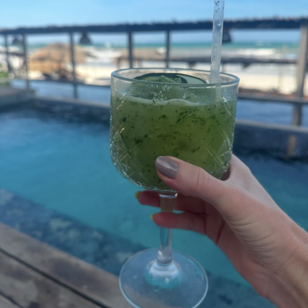 A woman's hand holding a glass with green juice in front of a pool and ocean