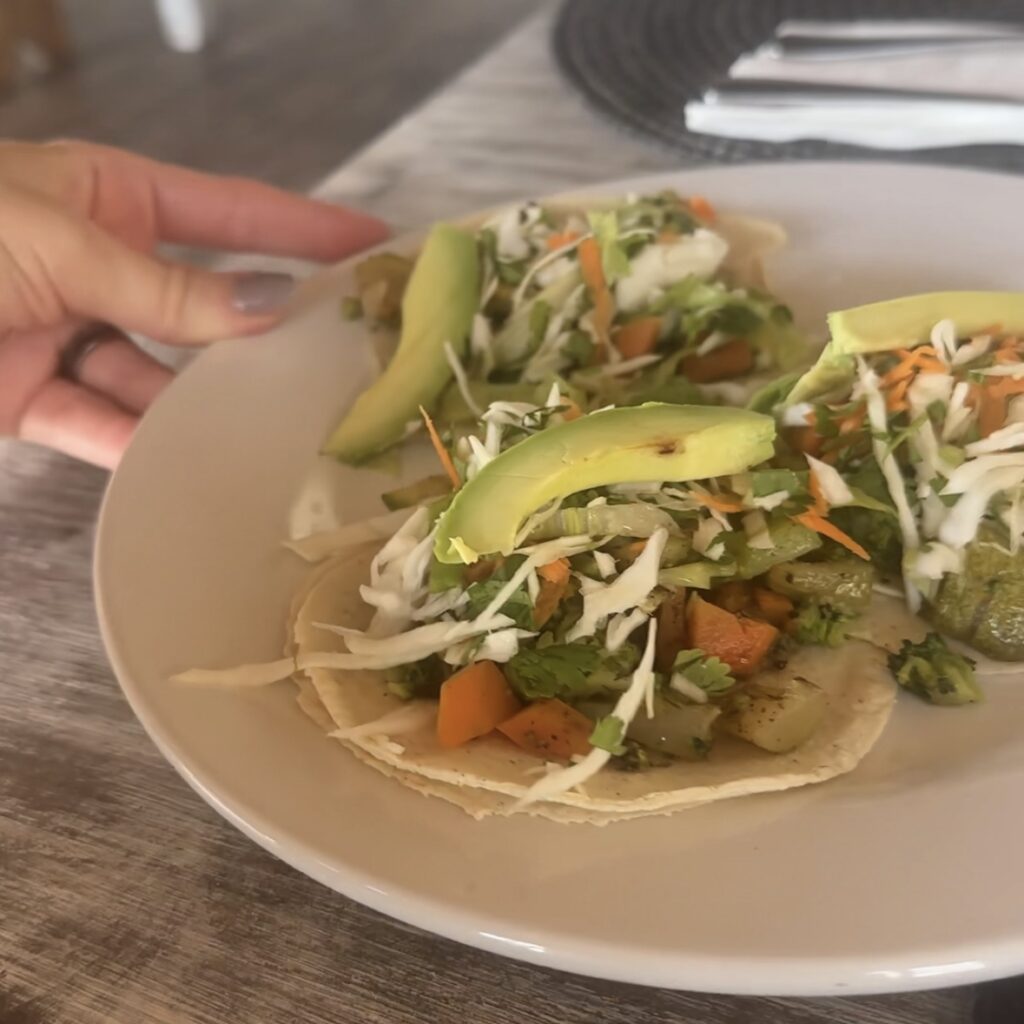 A woman's hand holding a white plate with 3 tacos with vegetables and avocado.