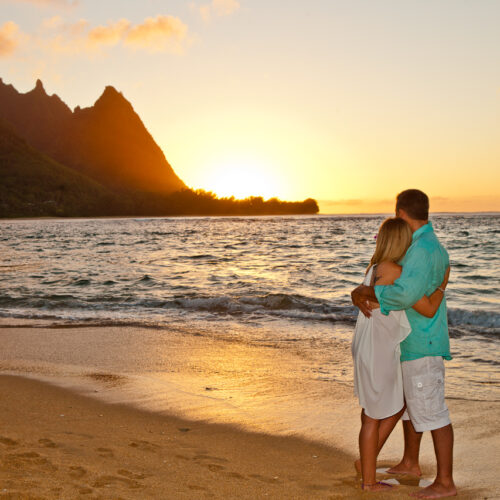 Ocean cliffs with a sunset in the background and a woman in a white dress with blonde hair and a man in a turquoise shirt and white shorts standing in the sand embracing