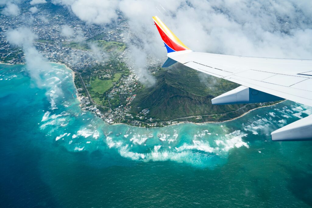 An airplane wing with yellow, red and blue on the wing looking down at a tropical island and clouds