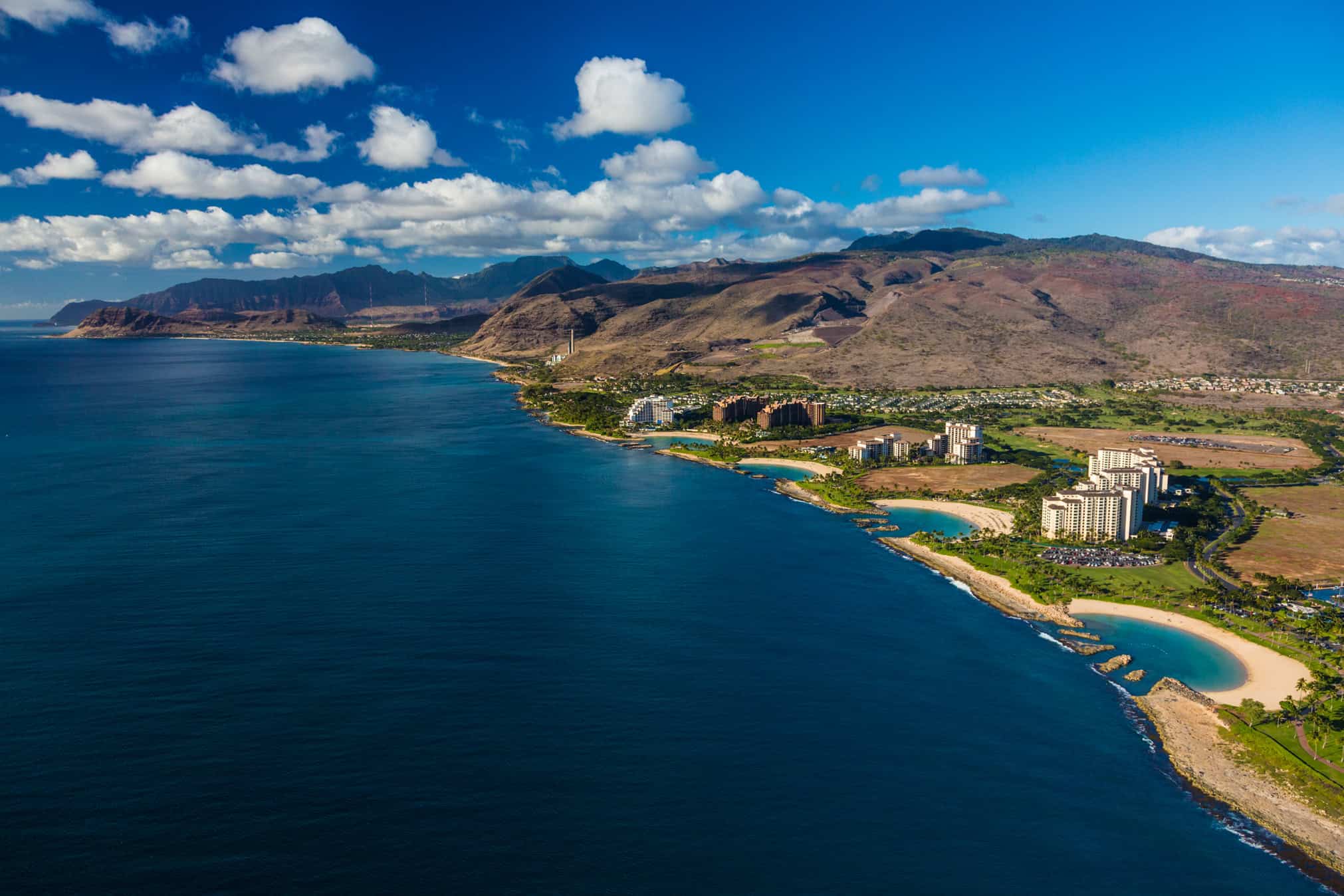 An aerial shot looking at a coastal town with dark blue water on the left and the coast to the right with mountains in the background.