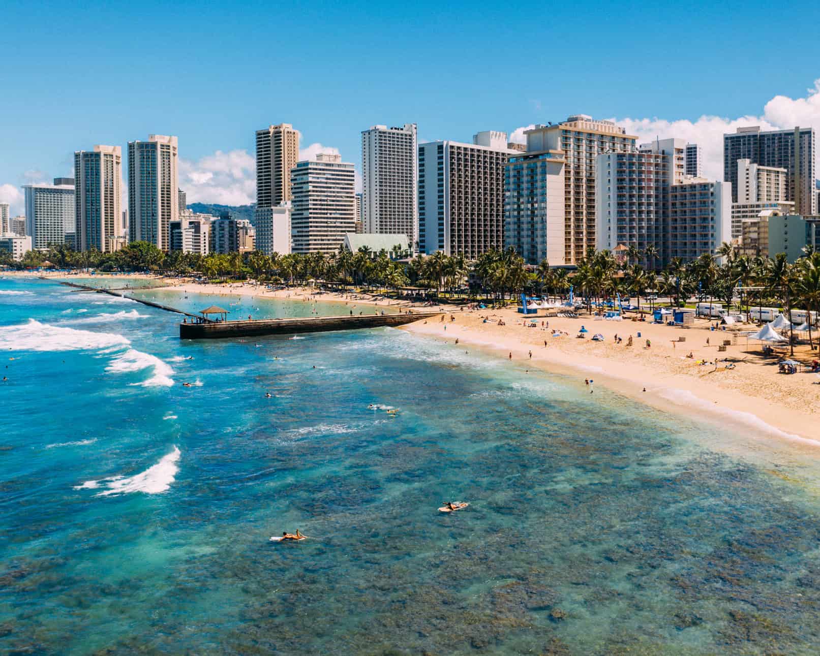 A city alongside a beach with many people on it and a dock jetting out into the ocean.