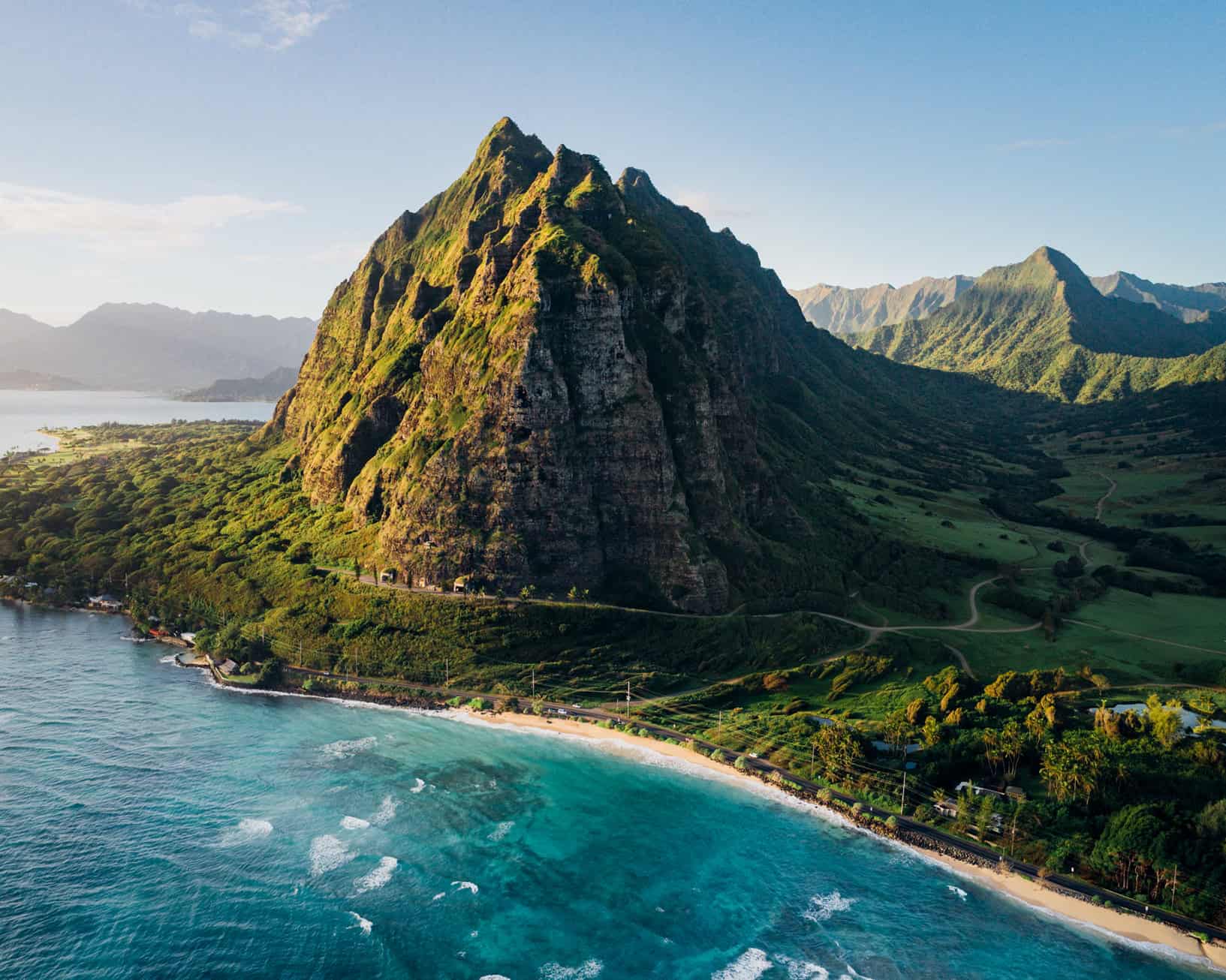 A tall green mountain surrounded by trees against a white sand beach and turquoise ocean.