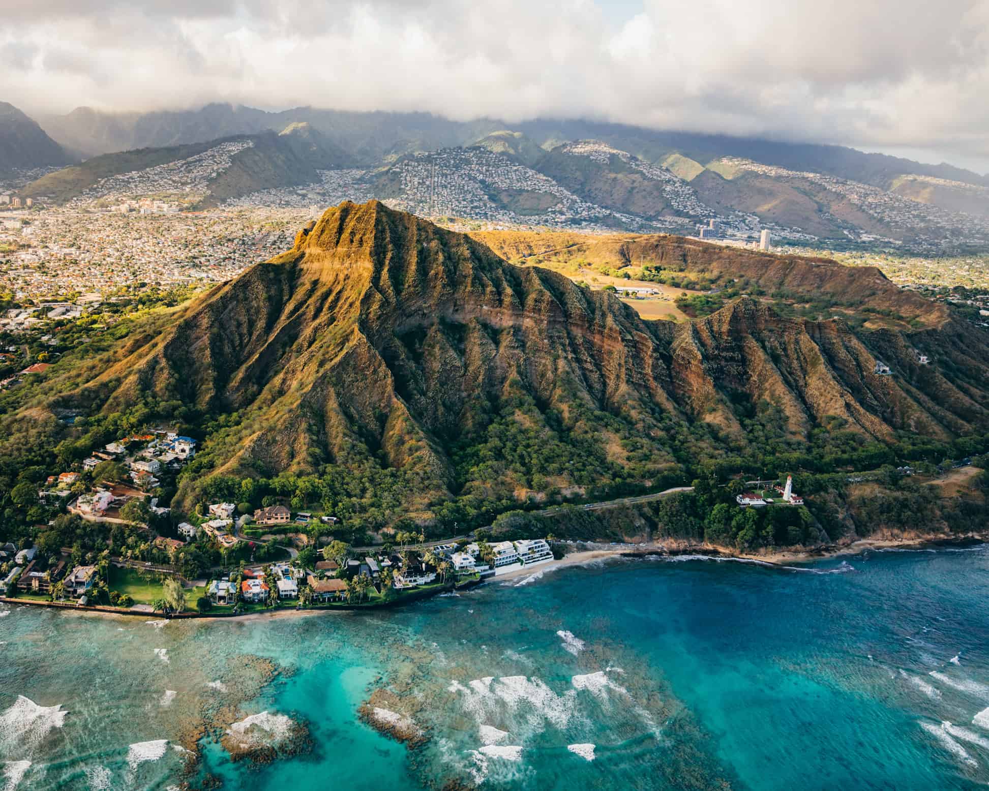 An aerial view of a mountain up against a turquoise ocean with coral in the water and roads and homes around the mountain