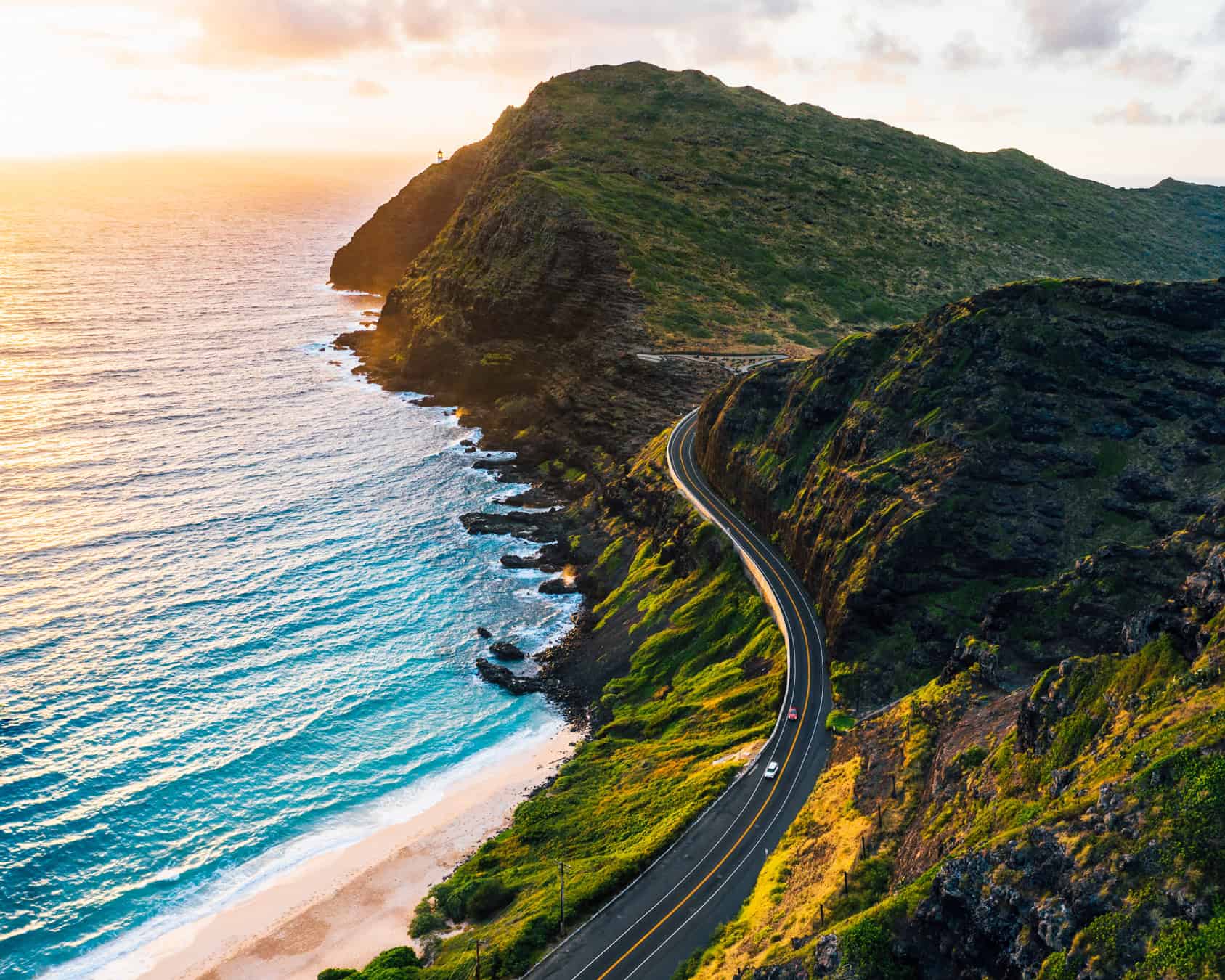 A winding road through mountains up against an ocean and beach with sun setting in the background.