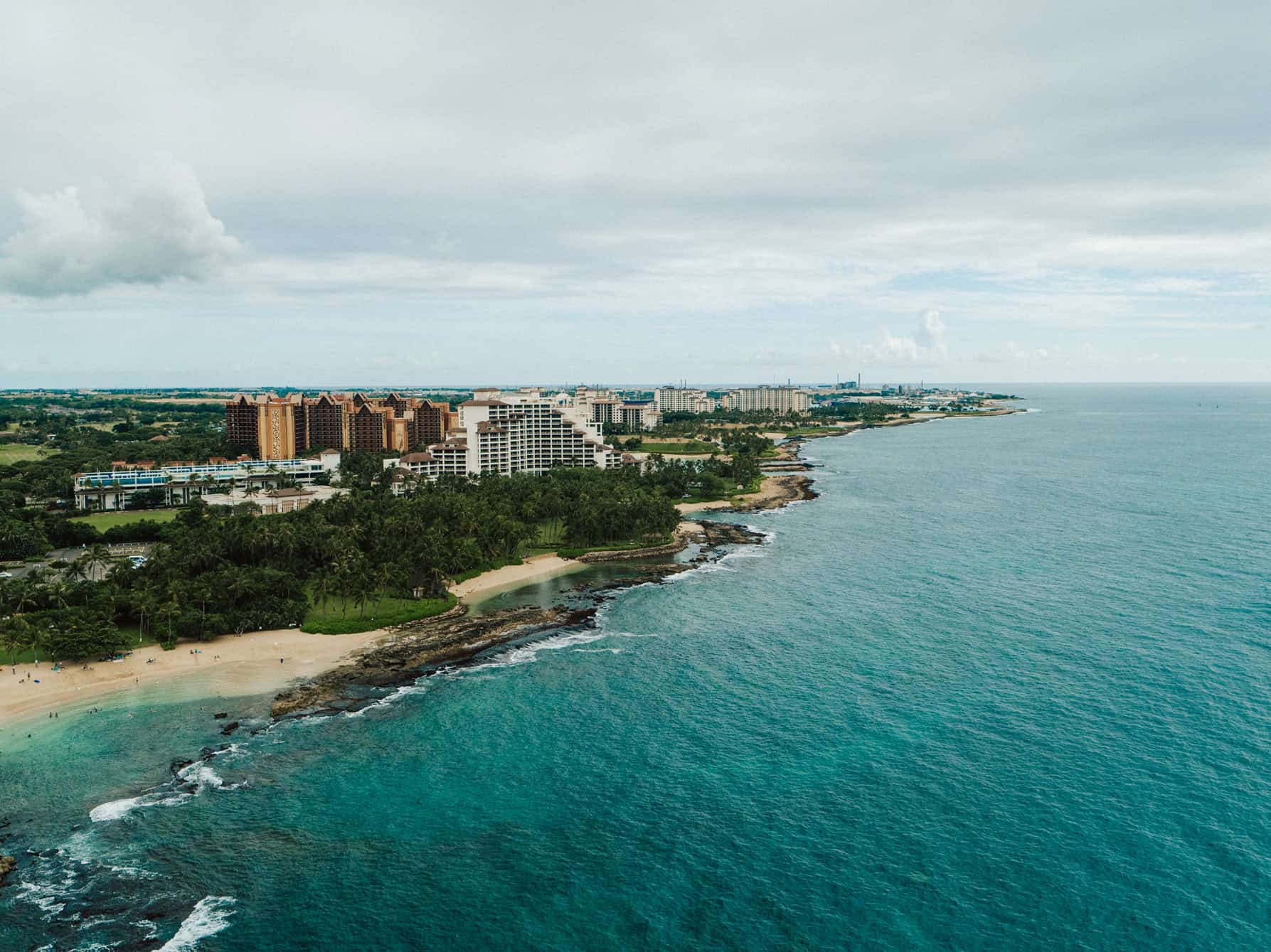 An aerial view over the ocean with hotels and tropical trees lining the shore.