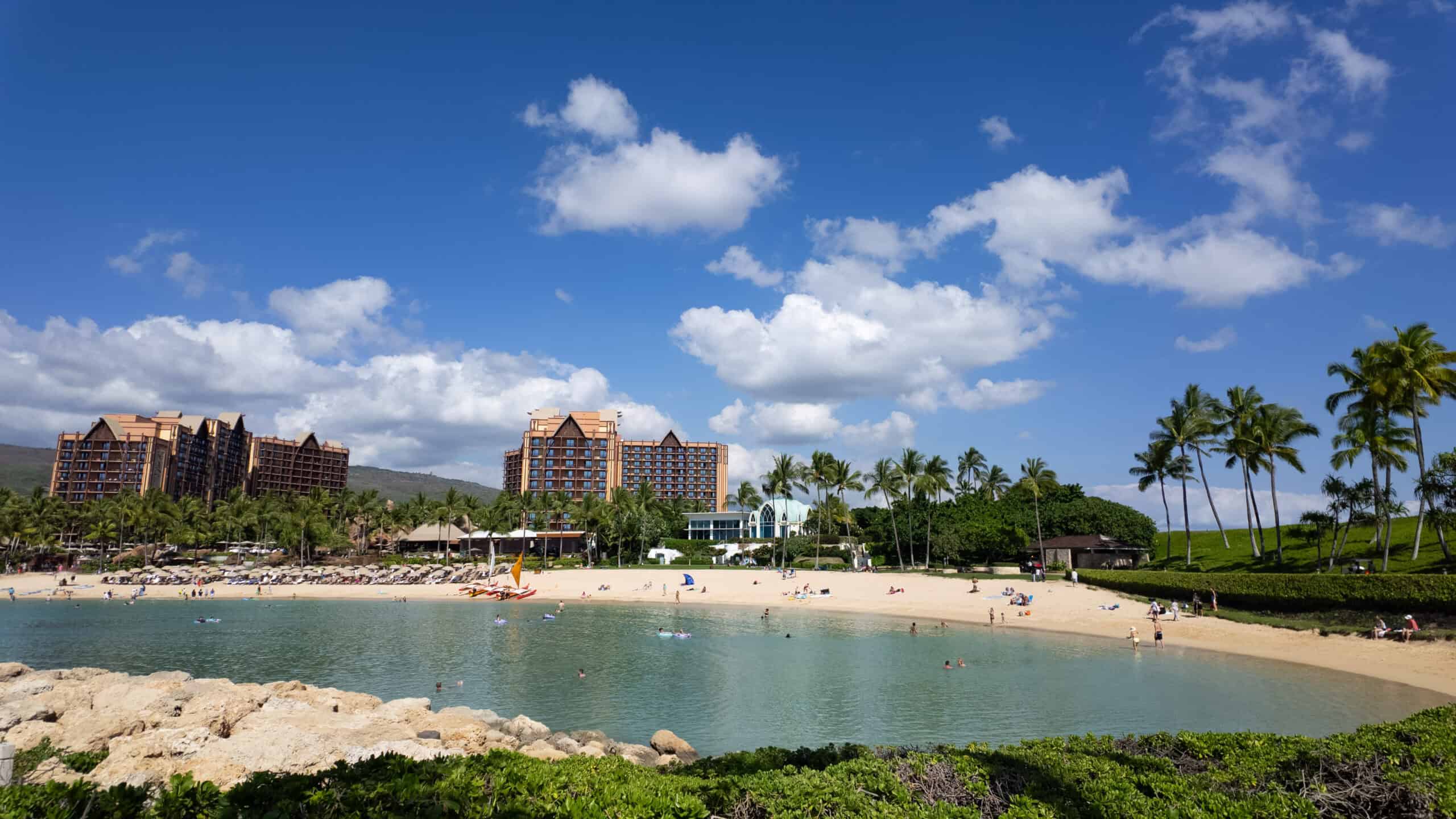 Looking out over a calm cove of water with beach-goers and a tan hotel with trees in the background.