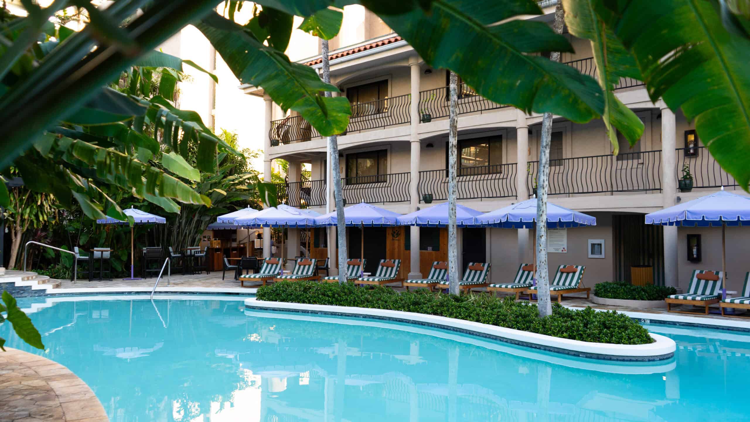 A calm pool of water lined with lounge chairs and purple umbrellas with a cream colored hotel in the background.