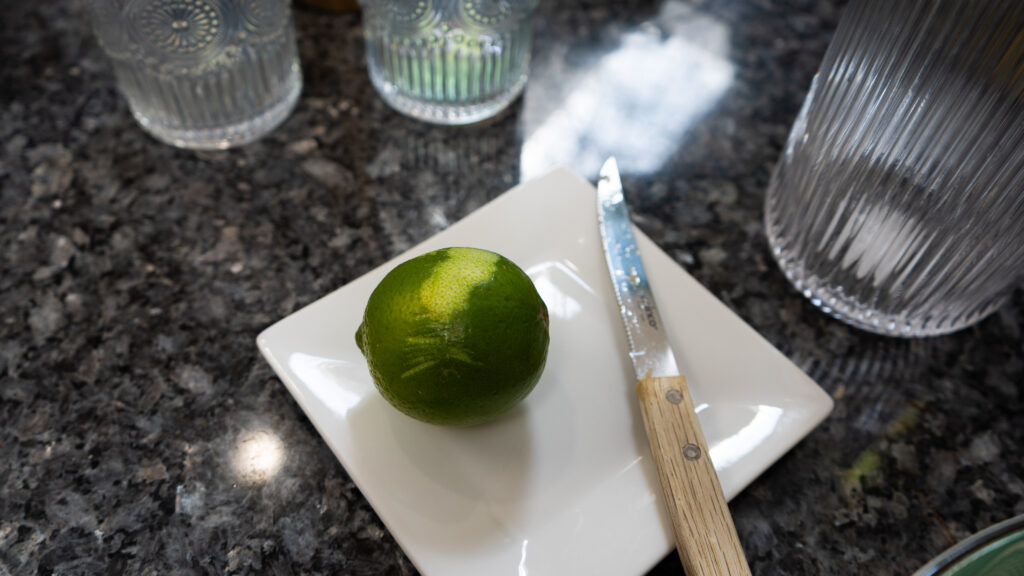 A lime on a white plate with a knife that has a wooden handle and two glasses to the side