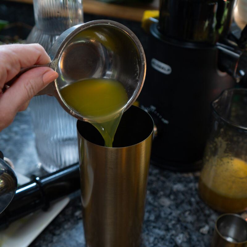 A yellow liquid being poured into a gold shaker with a juicer in the background