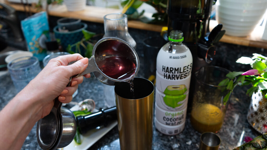 A hand pouring a pink liquid into a gold shaker with a bottle of Harmless Harvest coconut water to the side