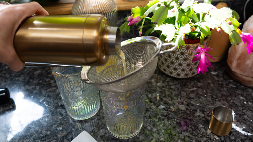 Pouring a yellow drink from a gold shaker into a glass cup with plants in the background.